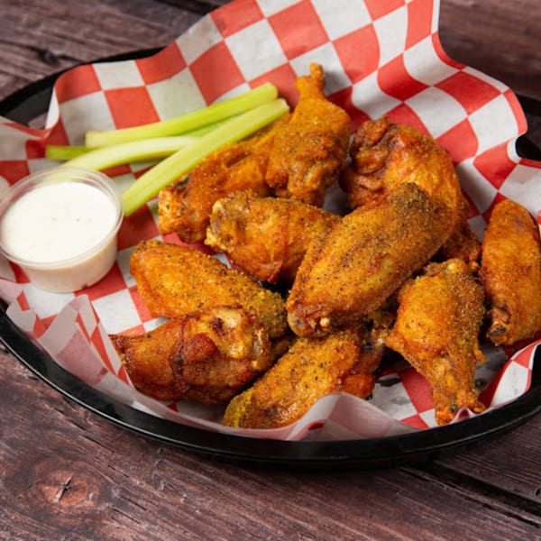 A black basket of Pop's Backdoor chicken wings, celery sticks, and ranch dressing on a wooden table.